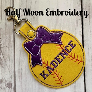 May include: Yellow softball-shaped leather keychain with a purple bow and the name "KADENCE" embroidered in black.