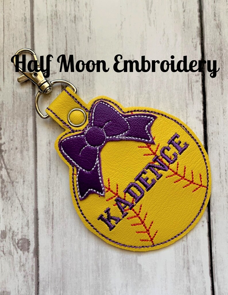 May include: Yellow softball-shaped leather keychain with a purple bow and the name "KADENCE" embroidered in black.