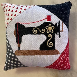May include: A white pillow with a black and gold embroidered sewing machine design. The pillow is made of patchwork fabric with red hearts, black and white gingham, and a red and white floral print.