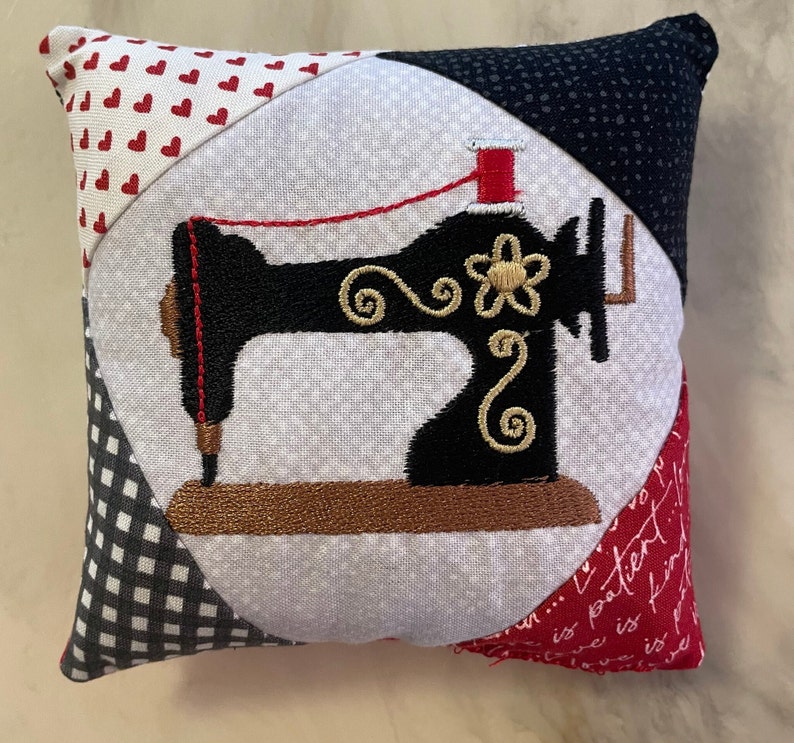 May include: A white pillow with a black and gold embroidered sewing machine design. The pillow is made of patchwork fabric with red hearts, black and white gingham, and a red and white floral print.