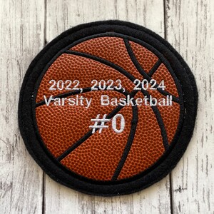 May include: A brown and black basketball with white stitching and the text "2022, 2023, 2024 Varsity Basketball #0" embroidered on it.