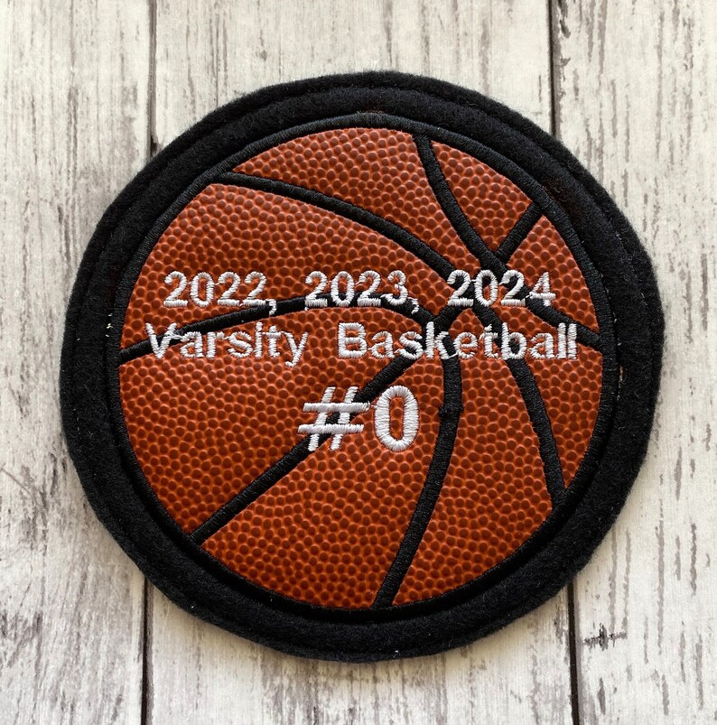 May include: A brown and black basketball with white stitching and the text "2022, 2023, 2024 Varsity Basketball #0" embroidered on it.
