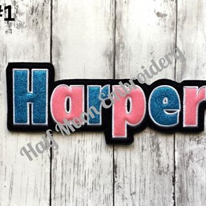 May include: A custom embroidered name patch featuring the name Harper in a bold, block-style font. The letters alternate between a shimmering blue glitter finish and a solid pink fabric, all outlined in white thread against a black felt border.