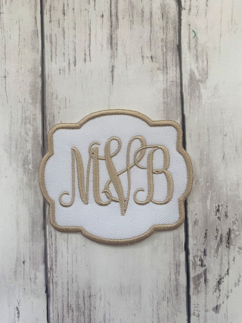 May include: White embroidered patch with a gold border and gold monogram letters "MVB". The patch has a decorative, scalloped edge. The background is a light wood grain.