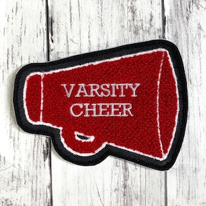 May include: A red megaphone-shaped patch with a black border and white stitching. The text "VARSITY CHEER" is embroidered in white letters across the center of the red megaphone. The patch has a textured surface and a distinct megaphone silhouette.