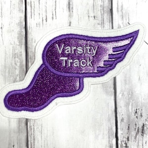 May include: A purple glittery fabric patch shaped like a winged foot with a white embroidered border. The text "Varsity Track" is stitched in silver thread across the center of the wing, featuring a sparkling texture throughout the design.