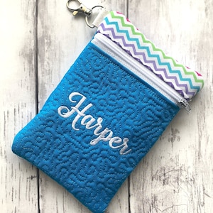May include: A blue zippered pouch with a white chevron pattern on the inside. The pouch has a white zipper and a white key chain loop. The pouch is embroidered with the name "Harper" in white.