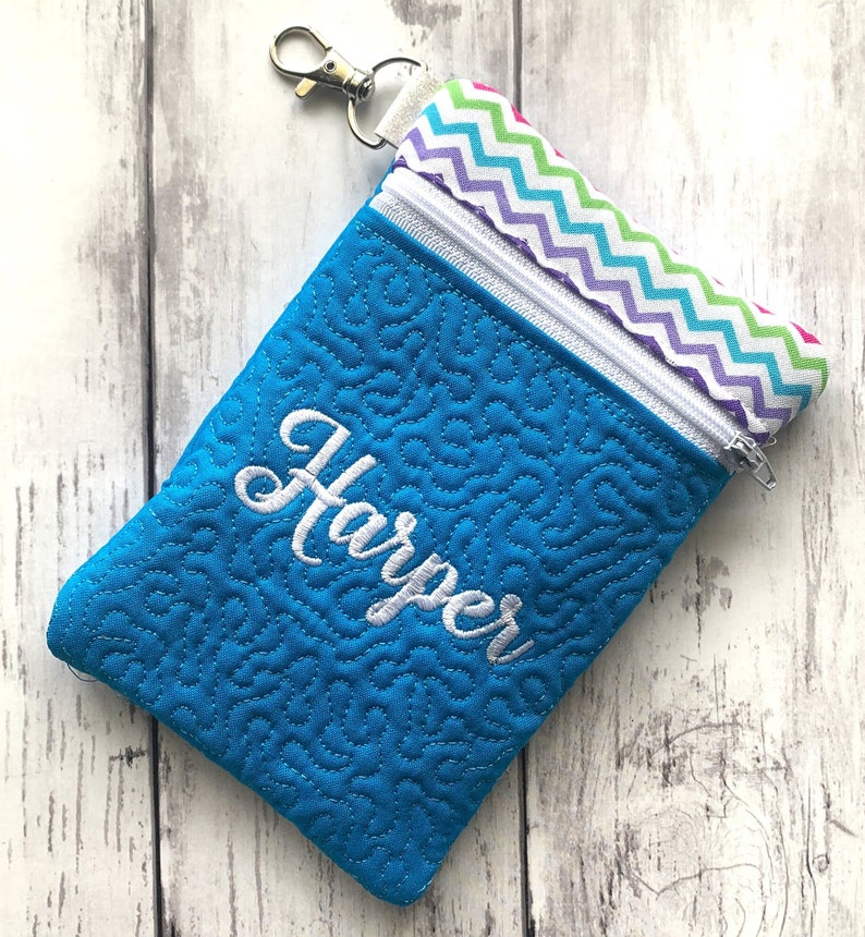 May include: A blue zippered pouch with a white chevron pattern on the inside. The pouch has a white zipper and a white key chain loop. The pouch is embroidered with the name "Harper" in white.