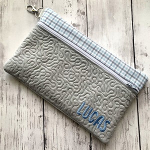 May include: A gray quilted zippered pouch with a blue and white plaid fabric lining. The pouch has a zipper closure and a silver loop for attaching a key chain. The pouch is personalized with the name "Lucas" embroidered in blue thread.