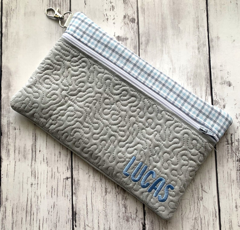 May include: A gray quilted zippered pouch with a blue and white plaid fabric lining. The pouch has a zipper closure and a silver loop for attaching a key chain. The pouch is personalized with the name "Lucas" embroidered in blue thread.