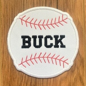 May include: White baseball-shaped patch with red stitching and the word "BUCK" in black lettering.