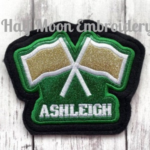 May include: Green and black embroidered patch with two gold and white crossed flags and the name "ASHLEIGH" in white lettering.