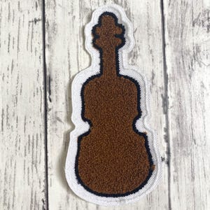 May include: A brown violin-shaped patch with a dark navy blue border and a white fabric backing. The surface of the violin shape has a textured, fuzzy appearance, and the entire patch is outlined with a white stitched edge.