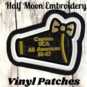 May include: A megaphone-shaped patch with black glitter vinyl, featuring gold embroidered text that reads Captain UCA All American 26-27. The patch has a gold glitter bow at the top and is bordered by thick white and black stitched layers.