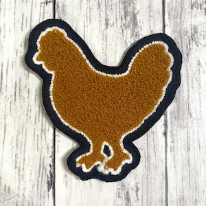May include: A punch needle patch shaped like a chicken, featuring a textured golden-brown body, a white outline, and a thick black border. The design is flat and shows the bird in profile, facing left with a distinct comb and tail feathers.