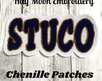 May include: A chenille patch with the word "STUCO" in navy blue letters with a gold outline, on a white background. The patch is in a wavy shape. The text "Half Moon Embroidery" and "Chenille Patches" are above and below the patch.
