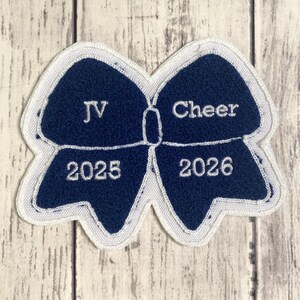 May include: A dark blue embroidered patch shaped like a bow with a white border. The left side features the text JV and 2025 in white thread, while the right side displays the text Cheer and 2026 in white thread, all set against a dark blue background.