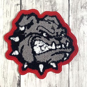 May include: A fabric patch featuring a stylized bulldog head in shades of gray, black, and white. The design is outlined with a thick red border, showcasing a textured, embroidered appearance with sharp teeth and a fierce expression on the dog.