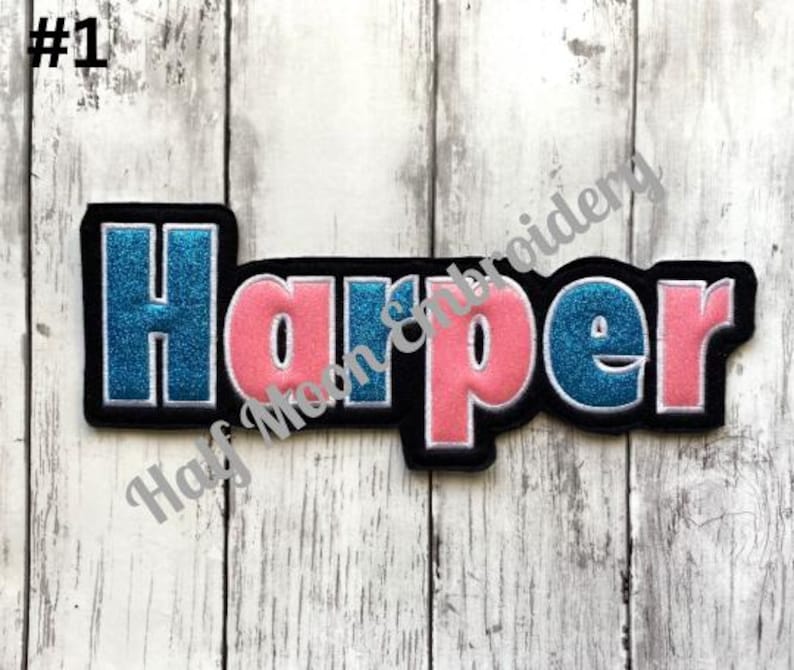 May include: A custom embroidered name patch featuring the name Harper in a bold, block-style font. The letters alternate between a shimmering blue glitter finish and a solid pink fabric, all outlined in white thread against a black felt border.