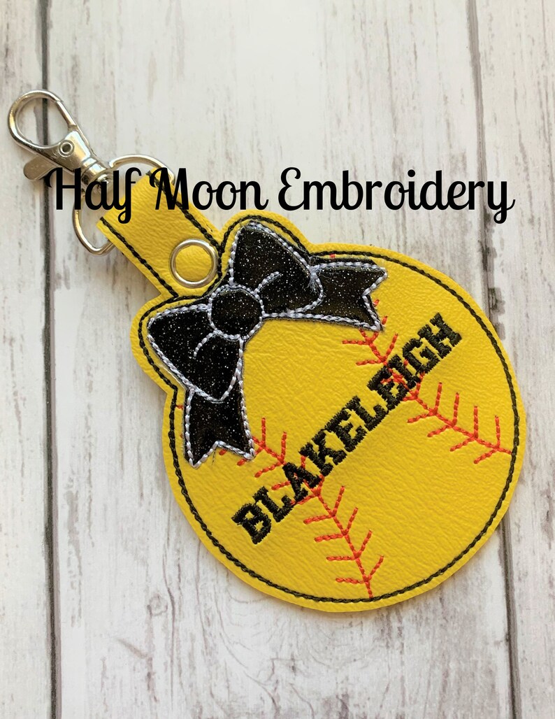 May include: Yellow softball keychain with black stitching and a black bow. The softball has the name "BLAKELY" embroidered on it.