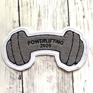May include: A gray embroidered patch in the shape of a barbell, with the text "POWERLIFTING 2025" in black. The patch has a white border and is likely for athletic apparel or accessories.