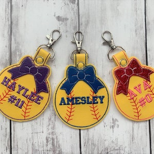 May include: Three yellow softball-shaped keychains with a bow and a name and number on each. The keychains are on a white wooden background. The names are Haylee #11, Aimesley, and Ava #0.