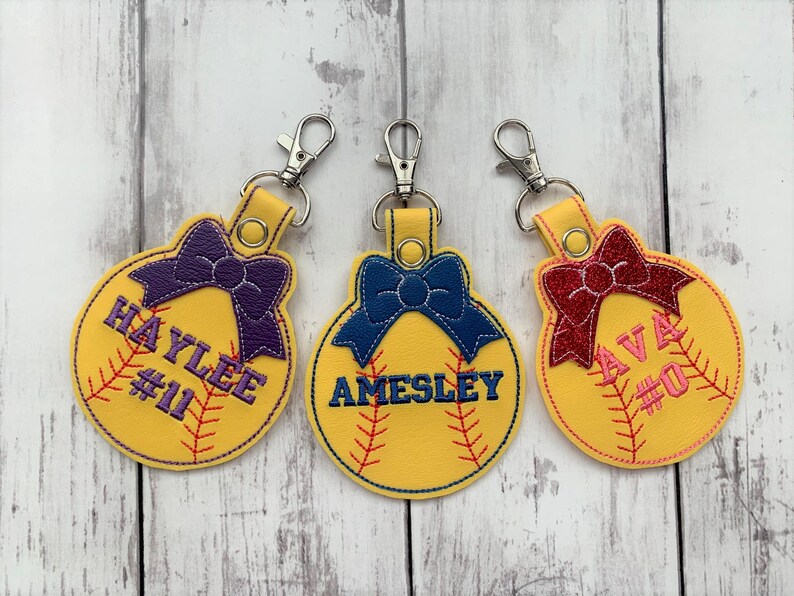 May include: Three yellow softball-shaped keychains with a bow and a name and number on each. The keychains are on a white wooden background. The names are Haylee #11, Aimesley, and Ava #0.