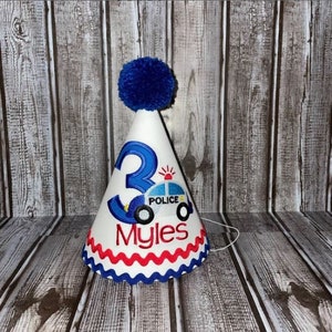 May include: White birthday party hat with a blue pom-pom on top. The hat has a blue number 3, a blue and white police car with the word "POLICE" on it, and the name "Myles" embroidered on it.