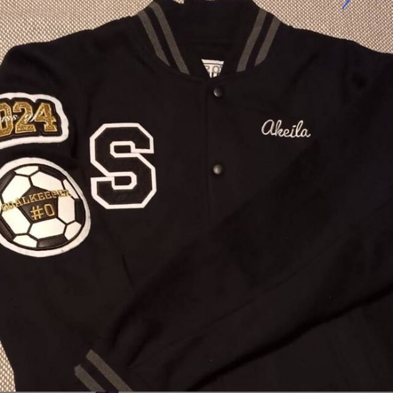K&ouml;nnte beinhalten: Schwarze Varsity-Jacke mit grauen Streifen an Kragen und &Auml;rmelb&uuml;ndchen. Die Jacke hat ein gro&szlig;es wei&szlig;es "S" auf der linken Seite und einen wei&szlig;en Fu&szlig;ball mit einer goldenen Kette und dem Text "Goalkeeper #0" auf dem linken &Auml;rmel. Die Jacke hat auch den Text "2024" in goldenen Glitzer auf der linken Schulter und den Namen "Akeila" in wei&szlig; gestickt auf der rechten Seite.