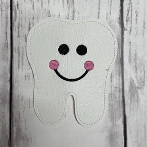 May include: A white tooth-shaped patch with a textured surface and stitched edges. The patch features an embroidered face with two round black eyes, two small pink circular cheeks, and a curved black line forming a simple smiling mouth.