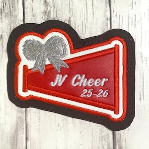May include: A red megaphone-shaped patch with a black border and white trim. It features a silver glitter bow at the top and white embroidered text that reads JV Cheer 25-26. The design is layered with a textured red surface and white stitching.