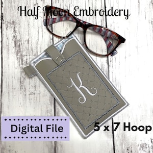 Pode incluir: Um estojo cinza acolchoado para óculos com um "K" bordado branco e uma borda prateada. O estojo tem um fecho de pressão e uma pequena alça. O texto "Half Moon Embroidery", "Digital File" e "5 x 7 Hoop" também é visível.