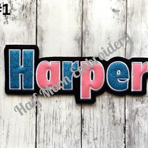 May include: A custom embroidered name patch featuring the name Harper in a bold, block-style font. The letters alternate between a shimmering blue glitter finish and a solid pink fabric, all outlined in white thread against a black felt border.