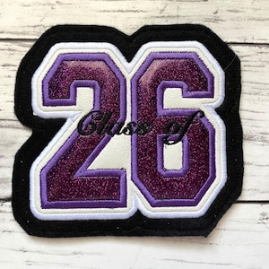 May include: A black, white, and purple glitter embroidered patch featuring the number 26 with the text Class of stitched in black cursive across the center. The patch has a thick black border and a white outline surrounding the purple numbers.