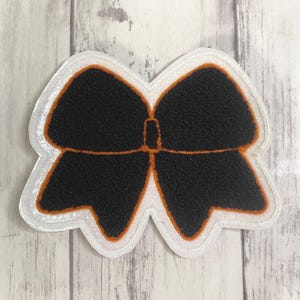 May include: A black bow-shaped patch featuring a textured surface with a thin orange outline. The patch is bordered by a wide, white, glittery fabric edge that follows the contour of the bow shape, creating a distinct layered appearance.