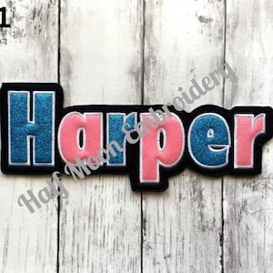 May include: A custom embroidered name patch featuring the name Harper in a bold, block-style font. The letters alternate between a shimmering blue glitter finish and a solid pink fabric, all outlined in white thread on a black felt background.