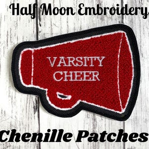 May include: A red megaphone-shaped chenille patch with a black border and white trim. The text "VARSITY CHEER" is embroidered in white across the center of the megaphone. The patch is displayed against a white wood-grain background.