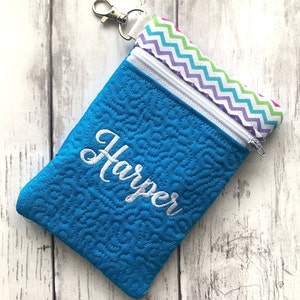 May include: A turquoise zippered pouch with a white quilted pattern and the name "Harper" embroidered on the front. The pouch has a rainbow chevron fabric lining and a silver zipper pull.