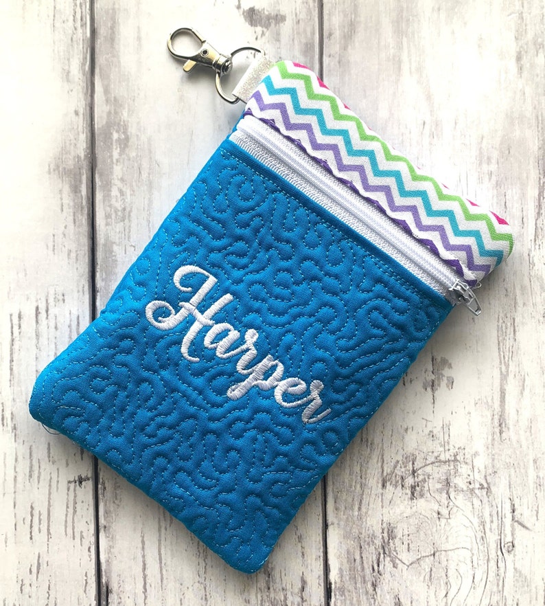 May include: A turquoise zippered pouch with a white quilted pattern and the name "Harper" embroidered on the front. The pouch has a rainbow chevron fabric lining and a silver zipper pull.