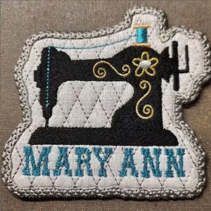 May include: A gray and white fabric patch with a black silhouette of a sewing machine. The machine has a gold thread and a gold flower design. The words "MARY ANN" are embroidered in blue below the machine.