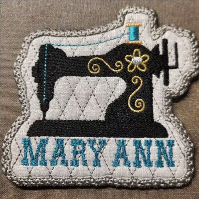 May include: A gray and white fabric patch with a black silhouette of a sewing machine. The machine has a gold thread and a gold flower design. The words "MARY ANN" are embroidered in blue below the machine.