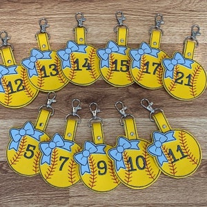 May include: Twelve yellow softball-shaped keychains with blue bows and black numbers 5, 7, 9, 10, 11, 12, 13, 14, 15, 17, 21.
