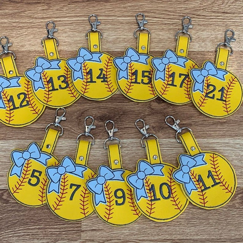 May include: Twelve yellow softball-shaped keychains with blue bows and black numbers 5, 7, 9, 10, 11, 12, 13, 14, 15, 17, 21.