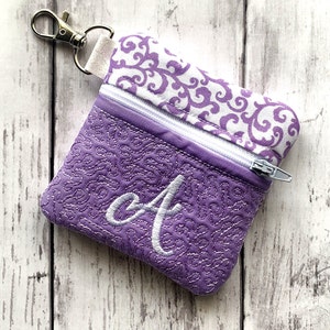 May include: A small purple zippered pouch with a white swirl pattern on the top flap. The pouch has a silver zipper and a silver key chain attached. The pouch is embroidered with a white letter "A" on the front.
