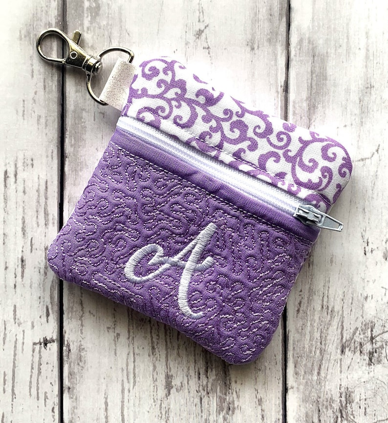 May include: A small purple zippered pouch with a white swirl pattern on the top flap. The pouch has a silver zipper and a silver key chain attached. The pouch is embroidered with a white letter "A" on the front.