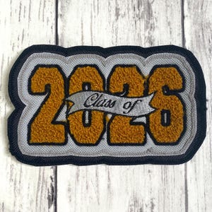 May include: A rectangular embroidered patch featuring the gold chenille numbers 2026 with a black border. A silver banner is stitched across the center with the black embroidered text Class of, set against a light gray background.