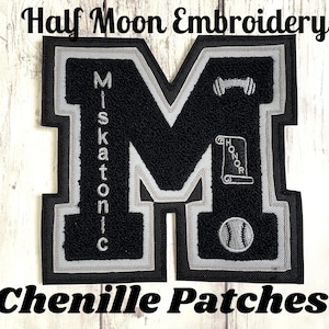 Puede incluir: Parche de chenilla negra con forma de la letra "M" con ribete gris y blanco. El parche presenta el texto "Miskatonic" verticalmente, y un icono de béisbol, pergamino y levantamiento de pesas. También se ve el texto "Half Moon Embroidery" y "Chenille Patches".