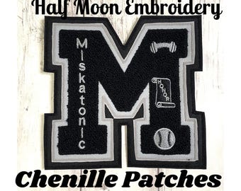 May include: A black and grey chenille letter M patch featuring the vertical text Miskatonic, a dumbbell icon, a scroll with the word Honor, and a baseball icon. The patch has a thick black border and a grey inner outline, with a textured surface.
