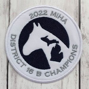 K&ouml;nnte beinhalten: Wei&szlig;er gestickter Aufn&auml;her mit einer schwarzen und wei&szlig;en Pferdesilhouette und dem Text "2022 MIHA District 16 B Champions" in silberner Schrift.