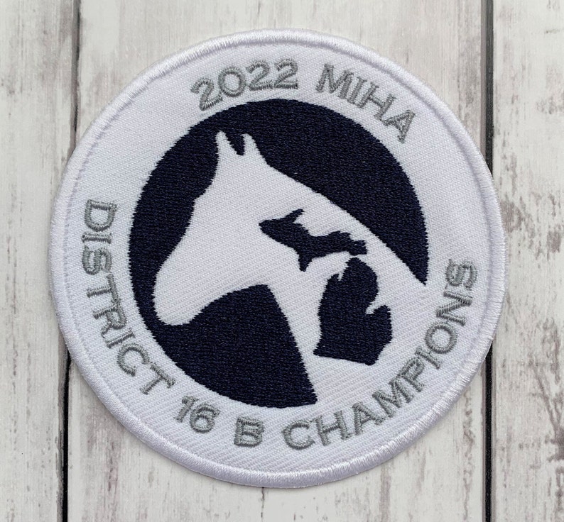 K&ouml;nnte beinhalten: Wei&szlig;er gestickter Aufn&auml;her mit einer schwarzen und wei&szlig;en Pferdesilhouette und dem Text "2022 MIHA District 16 B Champions" in silberner Schrift.